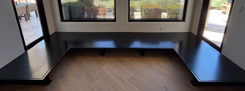 L-shaped black wooden bench installed along windows in a corner of a room with light wood flooring.