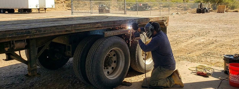 A person wearing protective gear welds near the wheels of a flatbed trailer outdoors, with equipment and tools visible nearby.
