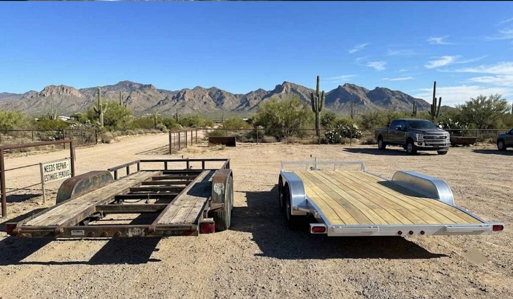 Two flatbed trailers are parked side by side on a dirt lot; one is old and rusted, the other is new and well-maintained, with mountains and desert vegetation in the background.