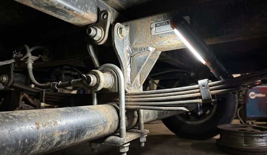 Close-up view of a vehicle's suspension system, showing leaf springs, bolts, brackets, and a flashlight illuminating the components.